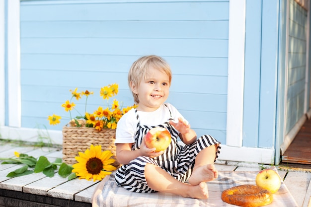 cute blond little boy with an apple and bun in his hands on porch of wooden house on summer day