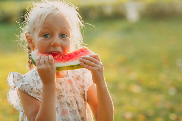 Cute blond little girl eats watermelon on the grass in park