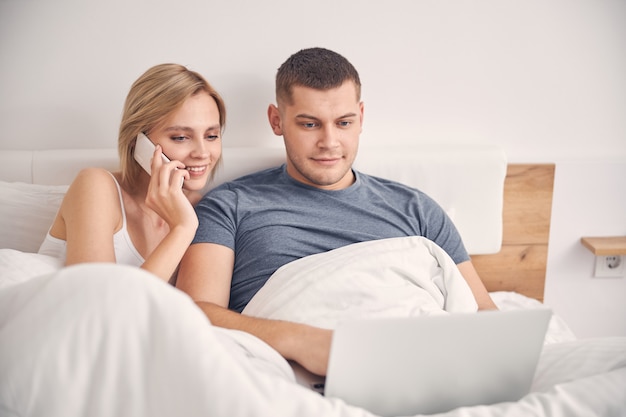 Cute blond woman talking on phone while blond man working on laptop in bed