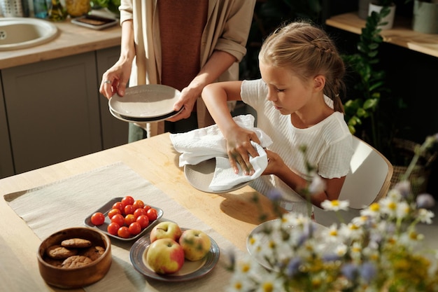 Cute blond youthful girl with white duster wiping clean plates after washing