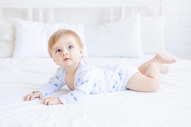 Cute blonde baby boy of six months is lying on his stomach on a big white bed at home and smiling the morning of a healthy baby