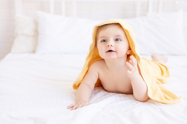 Cute blonde baby boy with a yellow towel on his head after a bath or bathing on a white bed at home in a bright room smiles the concept of child care and hygiene