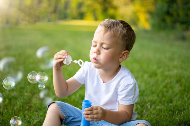 Carino bambino biondo gonfia bolle di sapone in estate su un prato verde, divertendosi, attività ricreative all'aperto