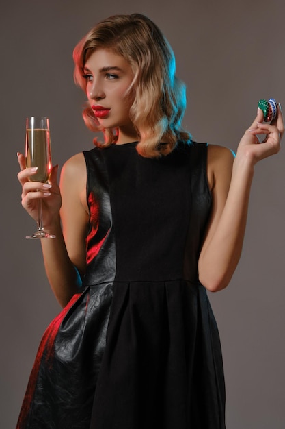 Cute blonde curly female bright makeup in black stylish dress holding glass of champagne and chips posing against gray studio background Gambling entertainment poker casino Closeup