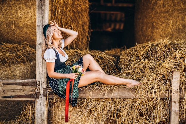 Cute blonde in dirndl, traditional festival dress with bouquet of field flowers sitting on the wooden fence at the farm