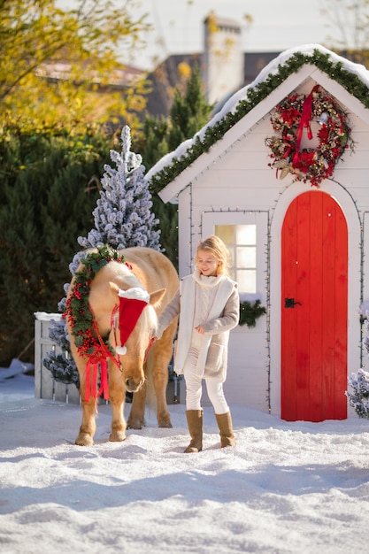 Cute blonde girl and adorable pony with wreath near the small house and snow-covered trees