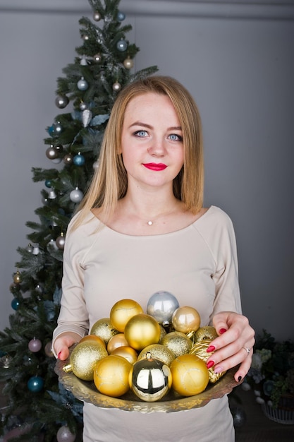 Cute blonde girl on beige dress against new year tree with with golden new year balls at studio Happy holidays