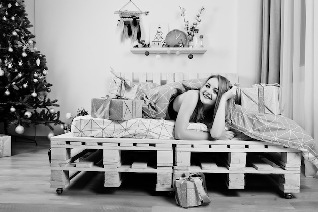 Photo cute blonde girl on black bra lying on bed with gifts boxes against new year tree at studio happy holidays