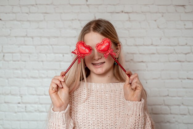 Ragazza bionda carina che copre gli occhi con cuori di tessuto rosso sul bastone