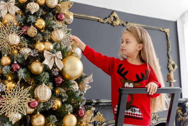 A cute blonde girl decorates a tree before Christmas at home. Happy childhood. Decorating a Christmas tree before Christmas.