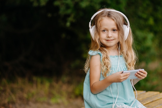 헤드폰에 귀여운 금발 소녀는 스마트 폰을 사용하고 밖에 앉아있는 동안 웃고 있습니다.