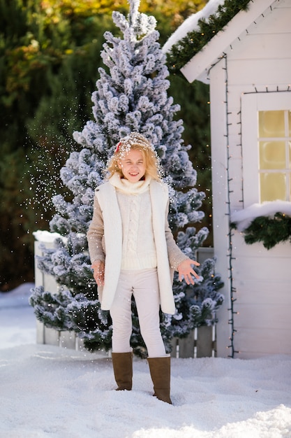 Cute blonde girl playing with snow near the small house and snow-covered trees