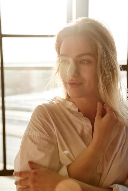 Cute blonde at home on the background of a window in a white men's shirt smiles tenderly and mysteriously look into the distance