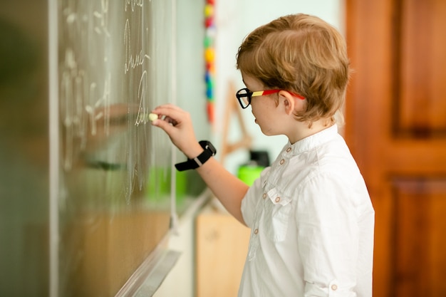 Cute blonde school student with stylish glasses writing in classroom