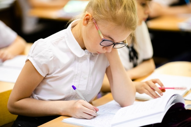 Foto studente di scuola biondo sveglio con i vetri alla moda che scrive nell'aula