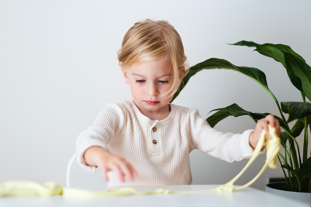 Cute blonde serious girltoddler playing with yellow sticky slime indoors at home on white backgroundEarly kid development messy game fun experiment concept