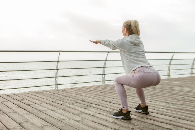 Cute blonde sportswoman trains on the beach. Young slim fit girl is practicing squats. Healthy lifestyle, fitness concept