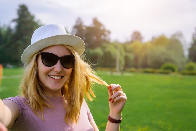 かわいいブロンドは、緑の夏の公園、休暇の概念でselfieを取ります。