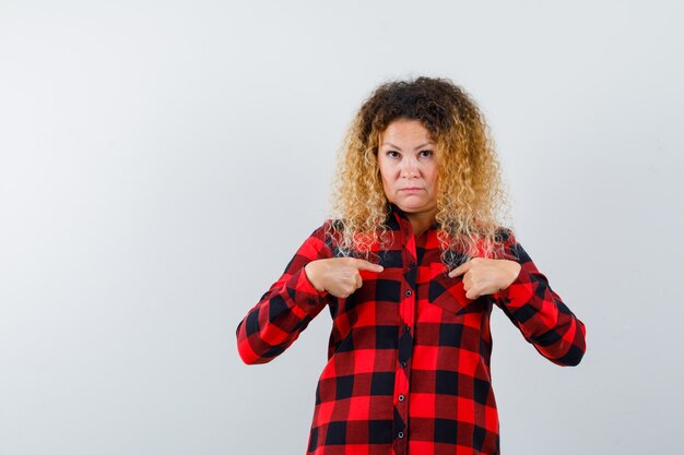 Photo cute blonde woman in checked shirt pointing at herself and looking confused , front view.