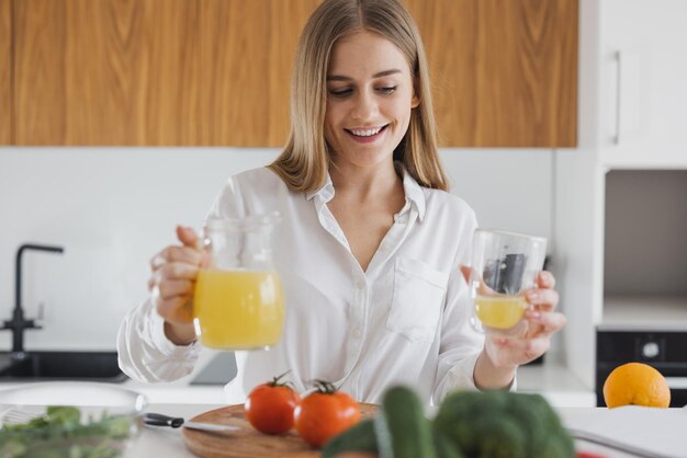 Photo cute blonde woman is looking at a recipe book and drinking juice in the kitchen