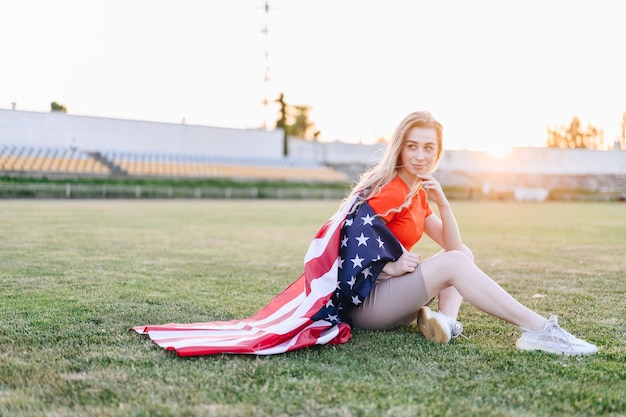 귀여운 금발의 여자는 경기장에서 녹색 잔디에 미국 국기에 싸여 앉아