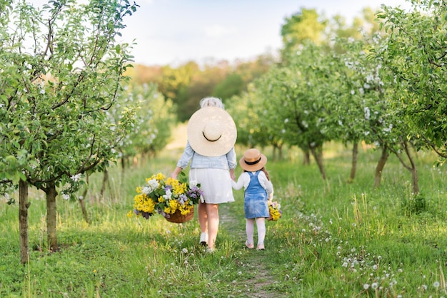 Cute blonde woman with her little daughter in a spring blooming garden They have big baskets of flowers and hats