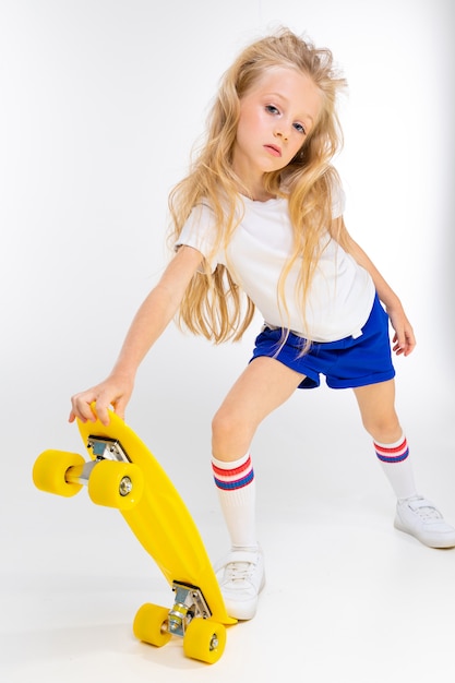 Cute blondy girl in sports shorts, a t-shirt and gym shoes with a yellow skateboard on a white wall