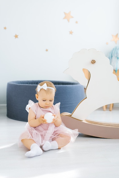 A cute blue-eyed one-year-old baby in a pink dress holds a small white ball in her hands and sits on the floor in the children's room