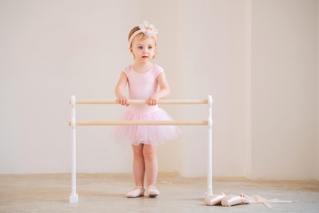 A cute blueeyed baby ballerina in a pink leotard and tutu standing near a ballet barre Dream concept of becoming a ballerina