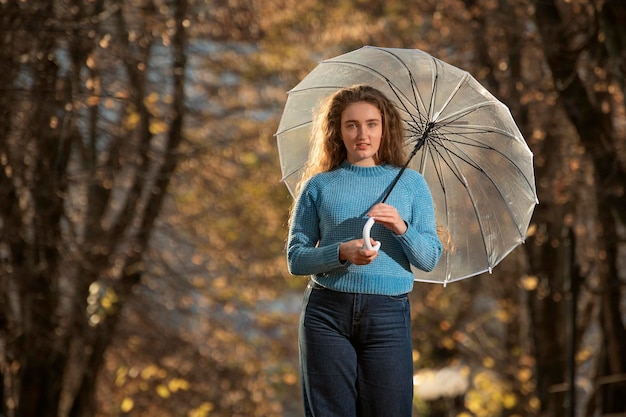 Cute blueeyed girl with wavy hair poses with transparent umbrella in hands against autumn forest