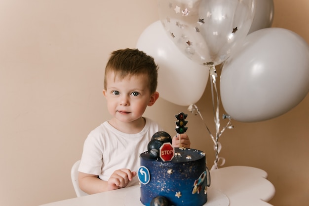 Il ragazzo carino di 3 anni festeggia il suo compleanno e mangia una deliziosa torta bellissima, foto di un bambino con palloncini