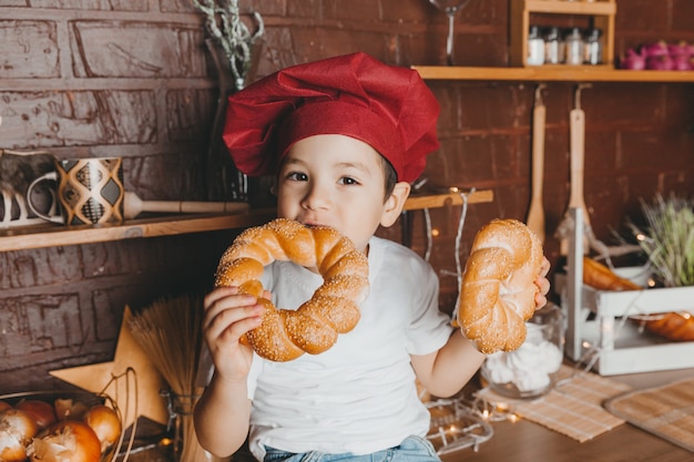 シェフの帽子をかぶったかわいい男の子がキッチンに座って、顔の近くにベーグルパンを持っています。