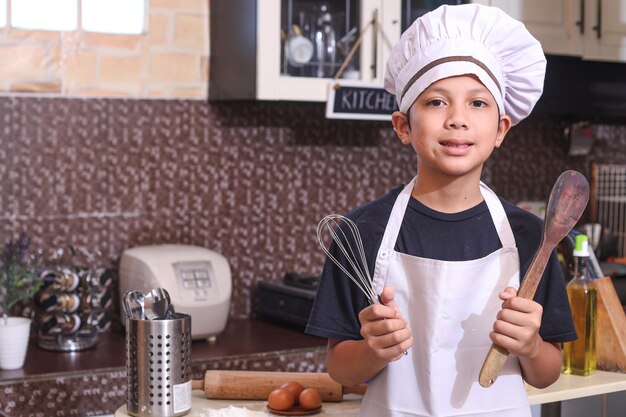 Cute boy in chef uniform holds wooden spatula and whisk against kitchen background