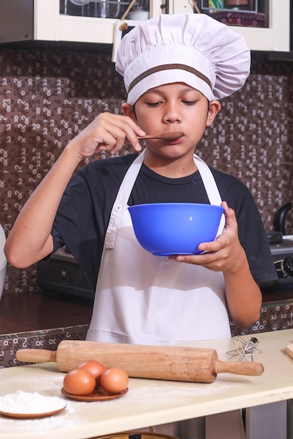 Cute boy in chef uniform taste the food on the bowl against kitchen background