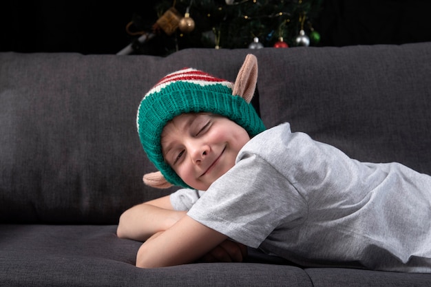 크리스마스 요정 모자를 쓴 귀여운 소년이 크리스마스 트리 배경에서 눈을 감고 소파에서 쉬고 있습니다. 휴일의 기대