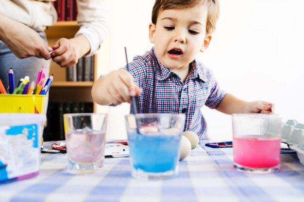 Cute boy coloring easter eggs on table against wall at home