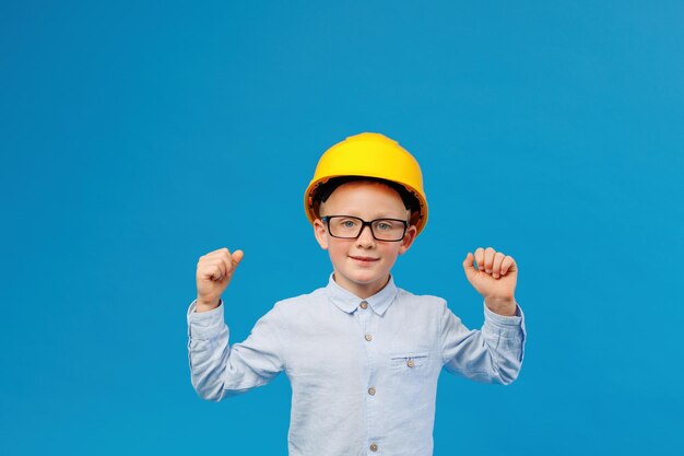 cute boy construction worker in a yellow hard hat stands in an indoor studio on a blue background and shows satisfaction with the work done
