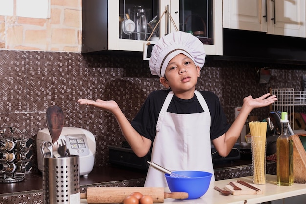 キッチンで料理をするかわいい男の子は、無知で混乱した表情で、腕と手を上げた。