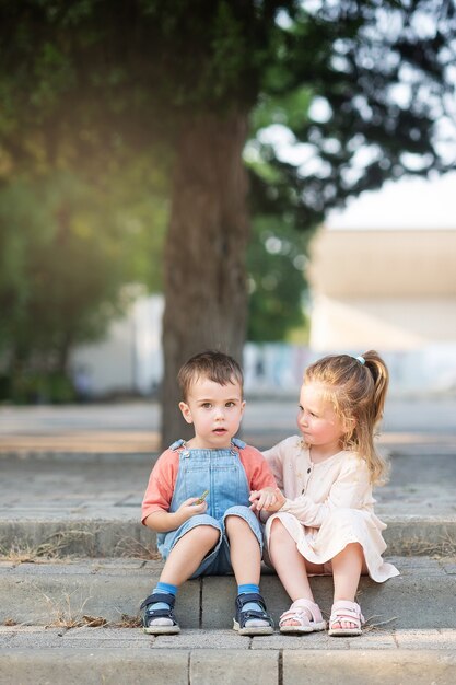 A  cute boy and a curly girl are sitting on a step under a tree in the park and looking at something