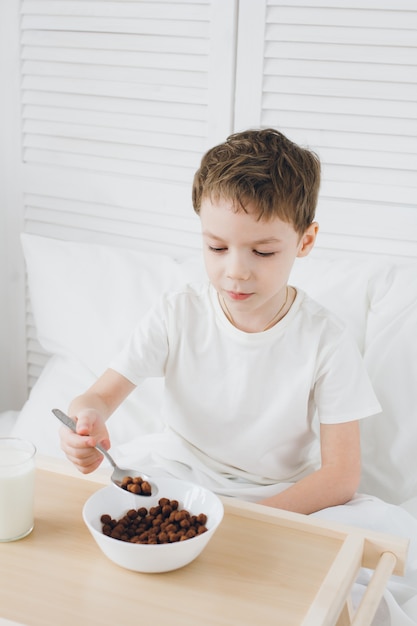 白いリネンとベッドに座って朝食チョコレートボールを食べるかわいい男の子