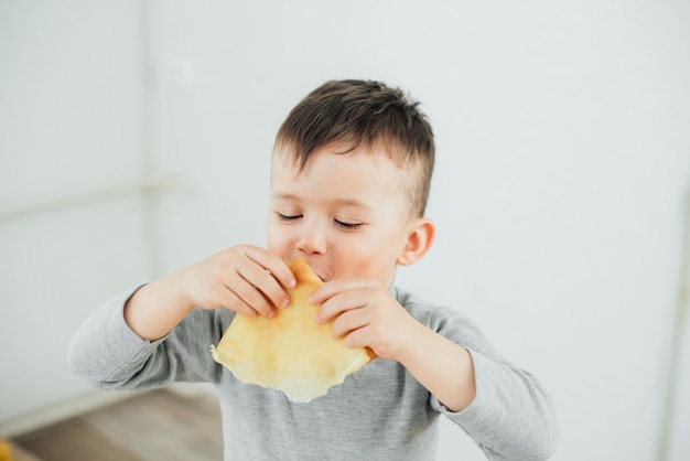 Cute boy eating delicious pancakes sitting at a table on a light background