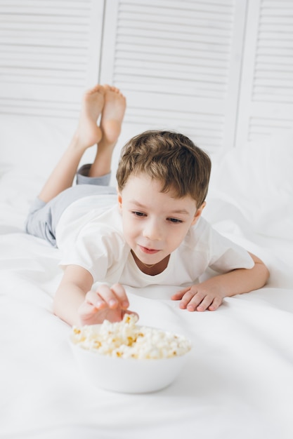 Милый мальчик ест попкорн, сидя в постели с белым постельным бельем