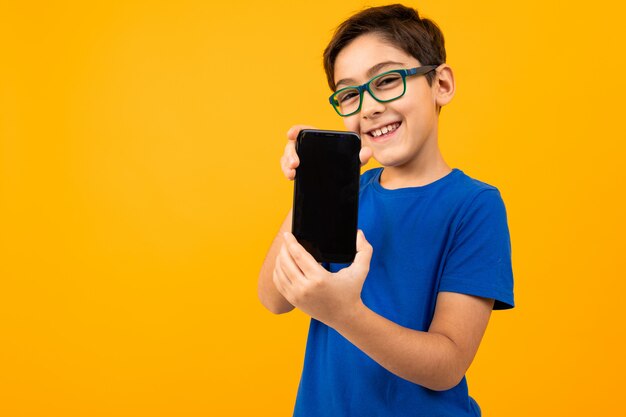 Милый мальчик в очках и синей футболке держит телефон с экраном вперед с макетом на желтом с копией пространства