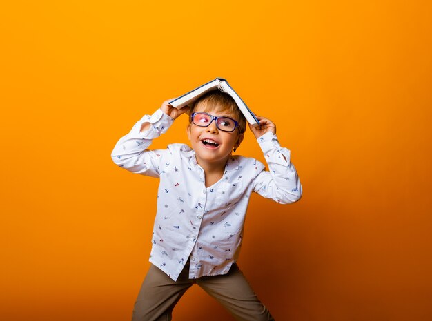 Cute boy in glasses with a book on his head shows tongue on yellow background