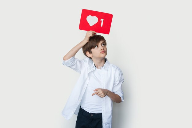 Photo cute boy is holding a paper with like sign above the head posing on a white studio wall