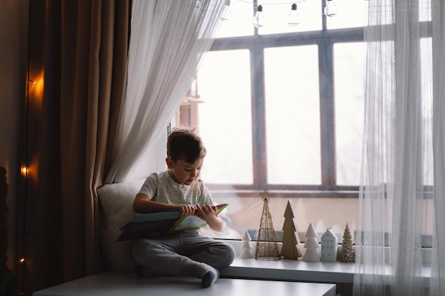 Cute boy is reading a book sitting on the windowsill near the window Cozy home with decorative Christmas trees Sweet home Winter holidays lifestyle