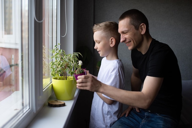 かわいい男の子が窓の近くで息子の隣にある屋内植物のお父さんに水をやっています