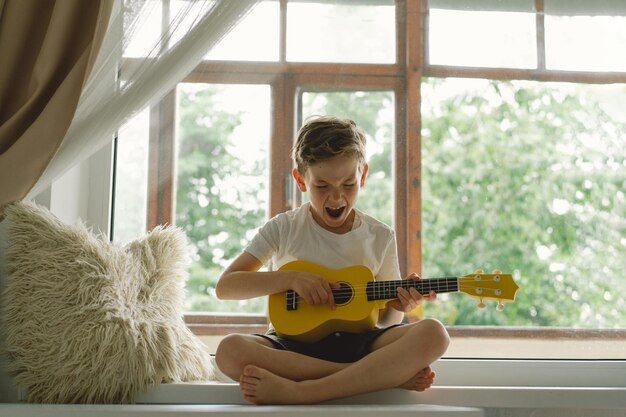 Cute boy learns to play the yellow Ukulele guitar on the windowsill near the window Cozy home Summer holidays lifestyle