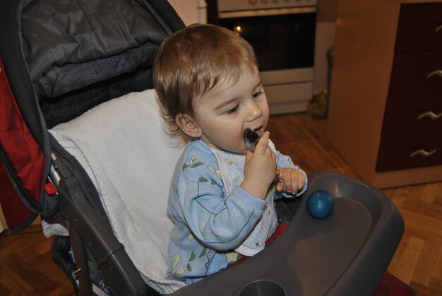 Photo cute boy licking spoon while sitting on baby stroller at home