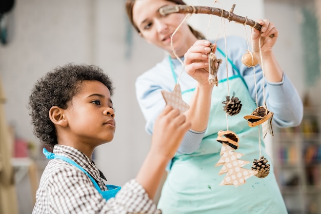 Cute boy looking at one of handmade Christmas decorations hanging on stick among pinecones and other toys